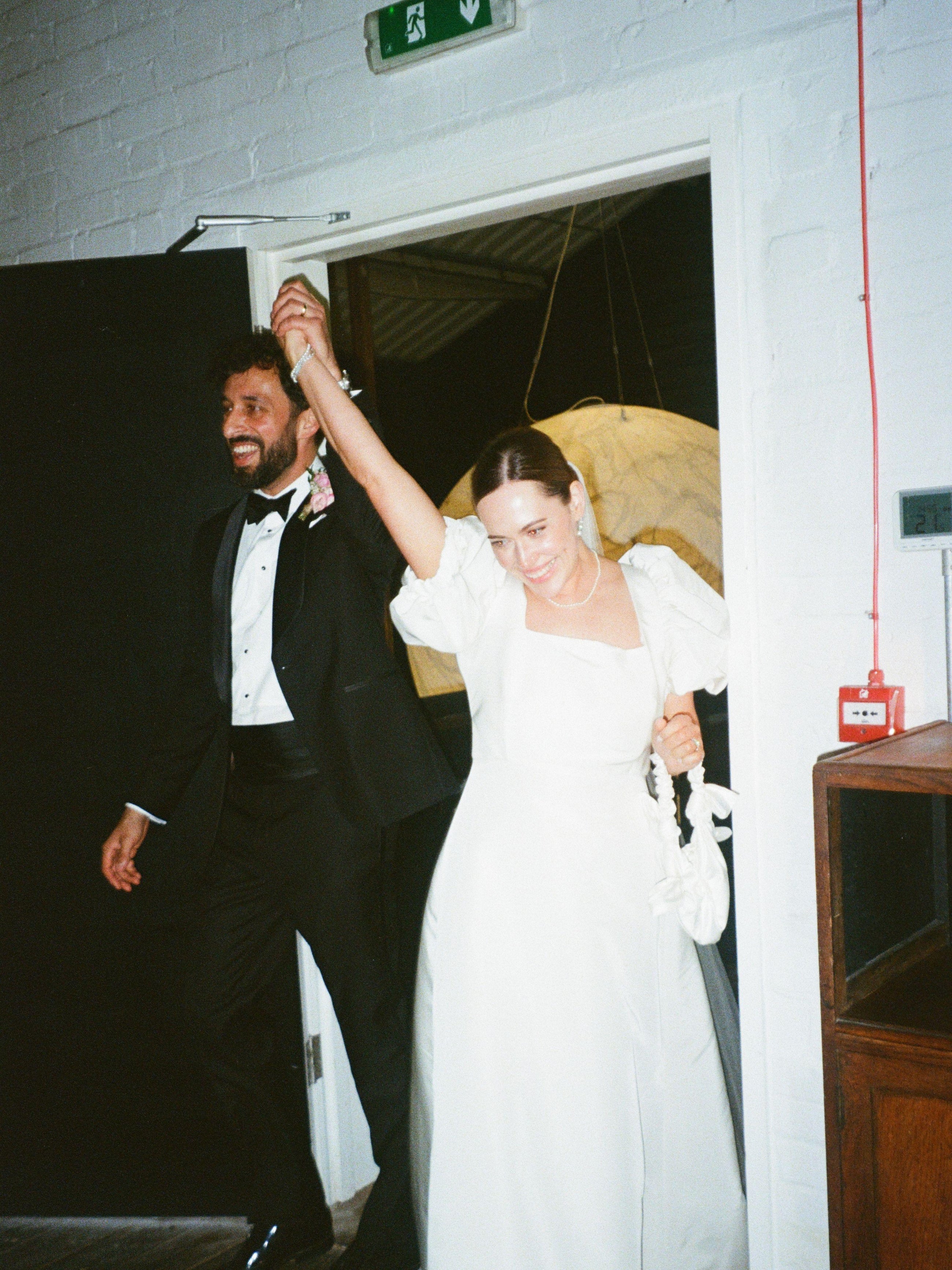 bride and groom enter a room happily. They are holding hands in the air, she holds a white satin knotted handbag