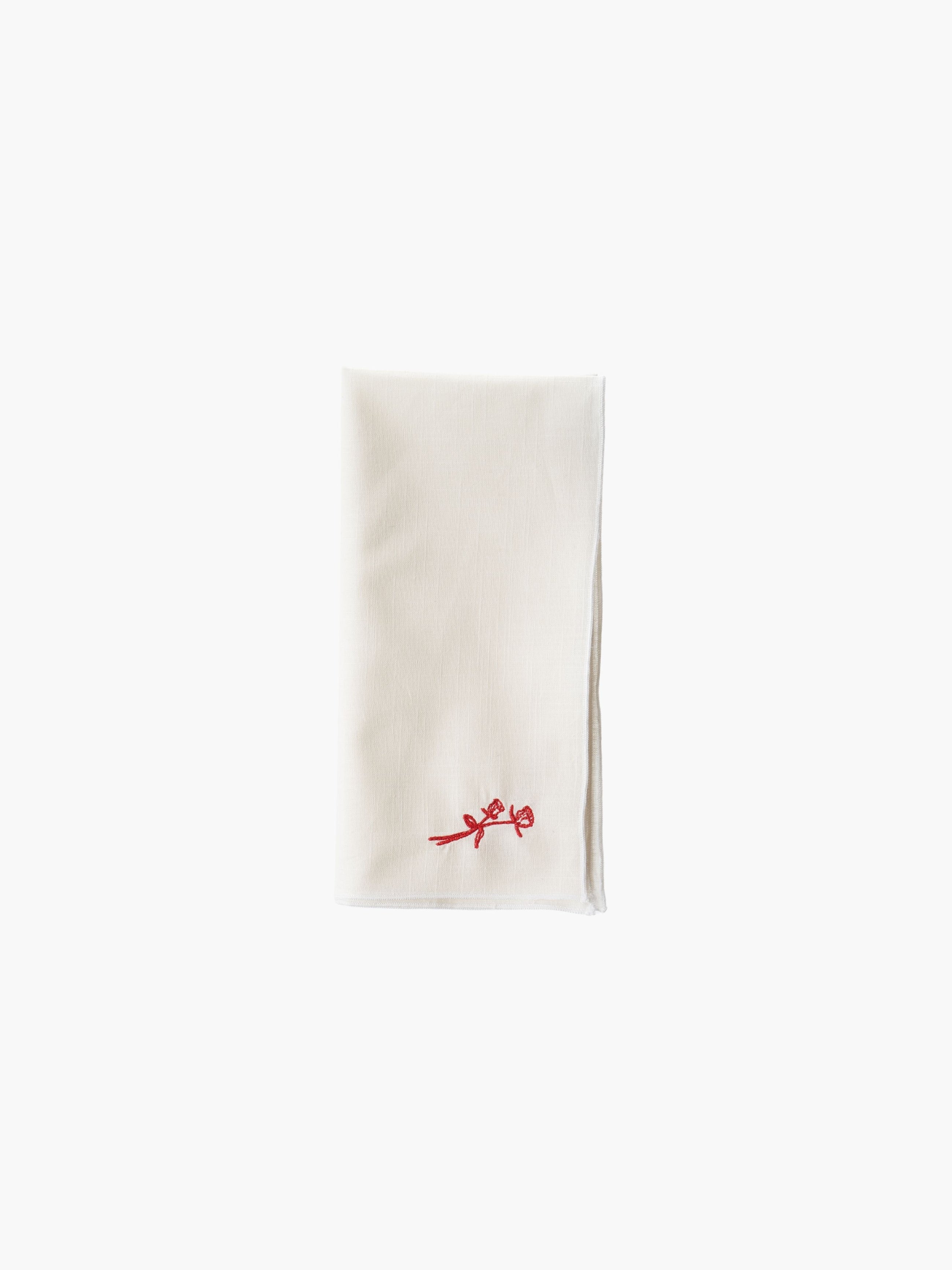 A folded cream-colored cloth napkin with a red embroidered rose design in the corner, set against a plain white background. Simple and elegant.
