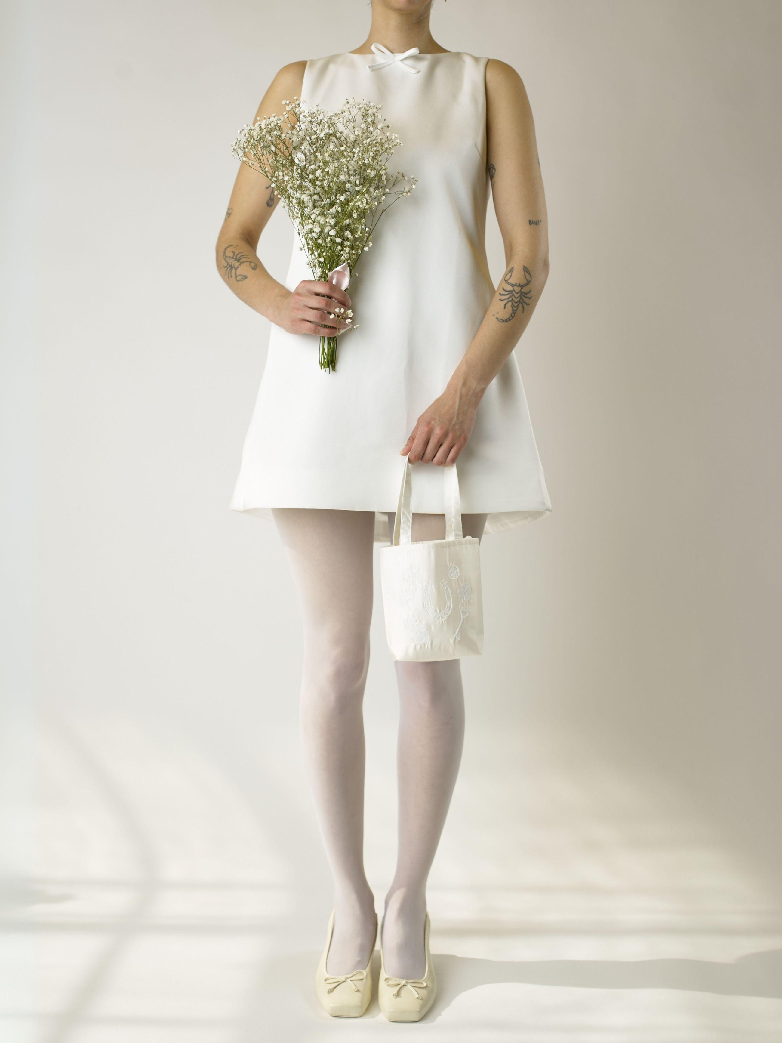 Person wearing a short ivory wedding dress, sheer white tights and ivory shoes, holding a bouquet of flowers and an ivory satin mini tote bag. Set against a plain background.
