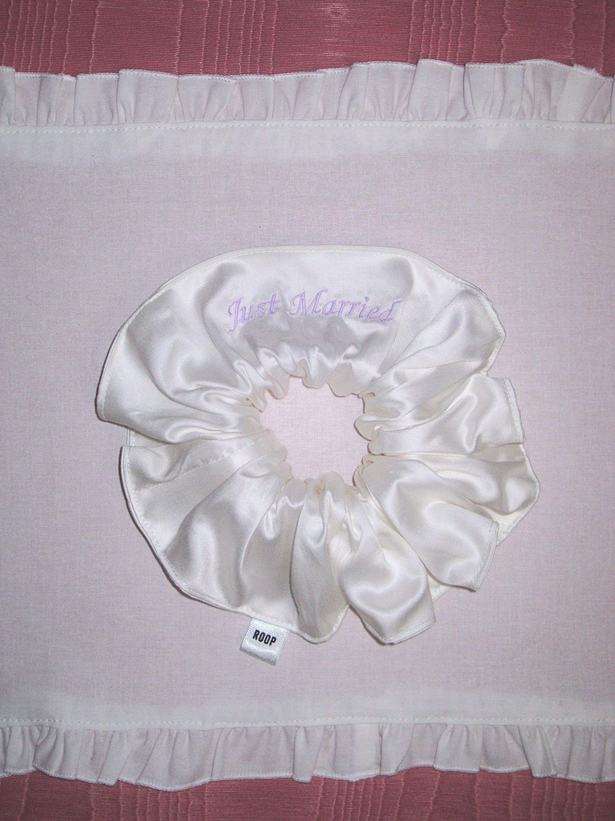 Ivory bamboo silk satin scrunchie with 'Just Married' embroidered in lilac on a white ruffled placemat, set on a pink moire background.