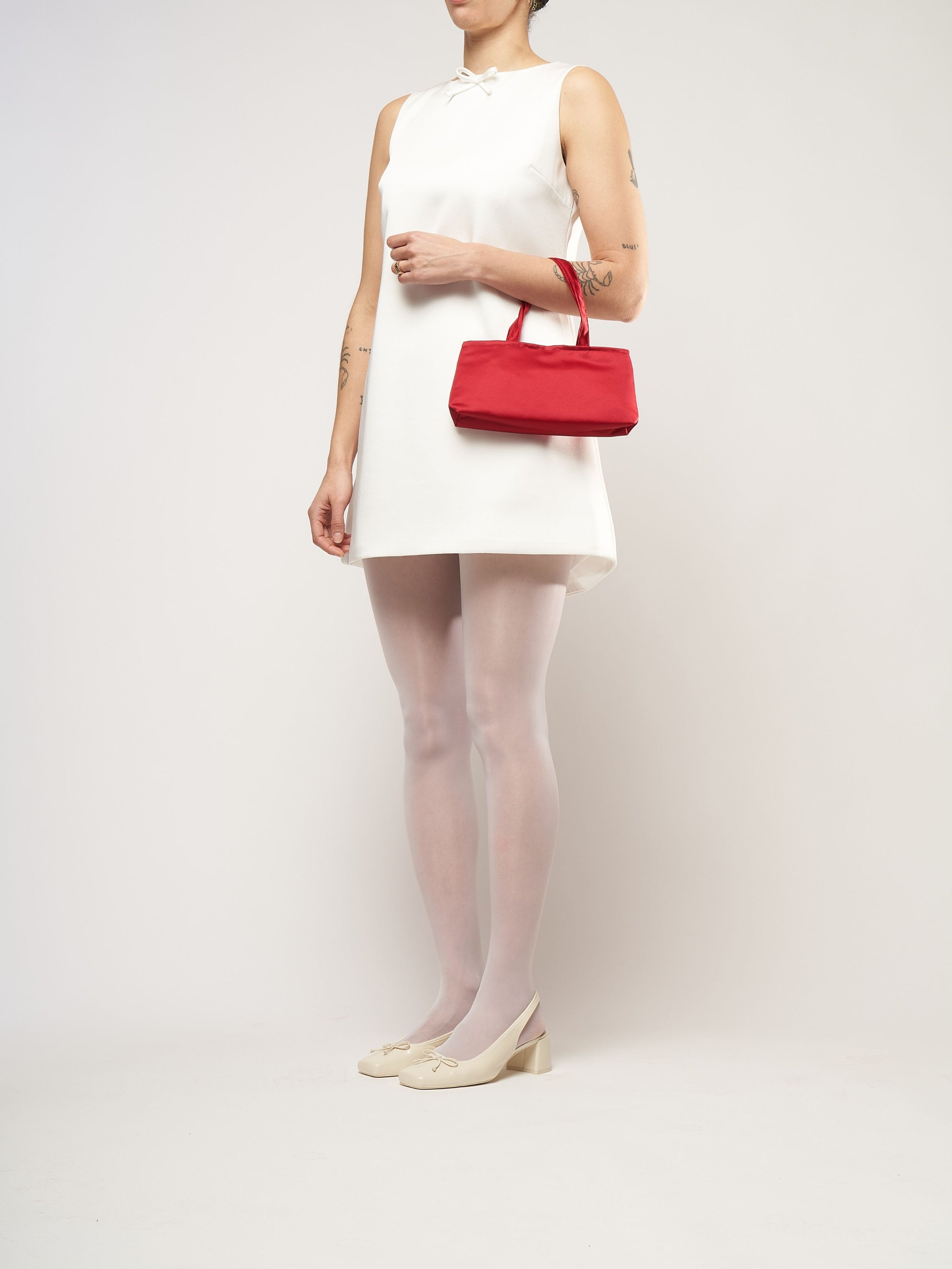 A woman in a sleeveless white dress holds a red handbag. She wears white tights, cream heels, and has tattoos on her arms, standing against a plain background.