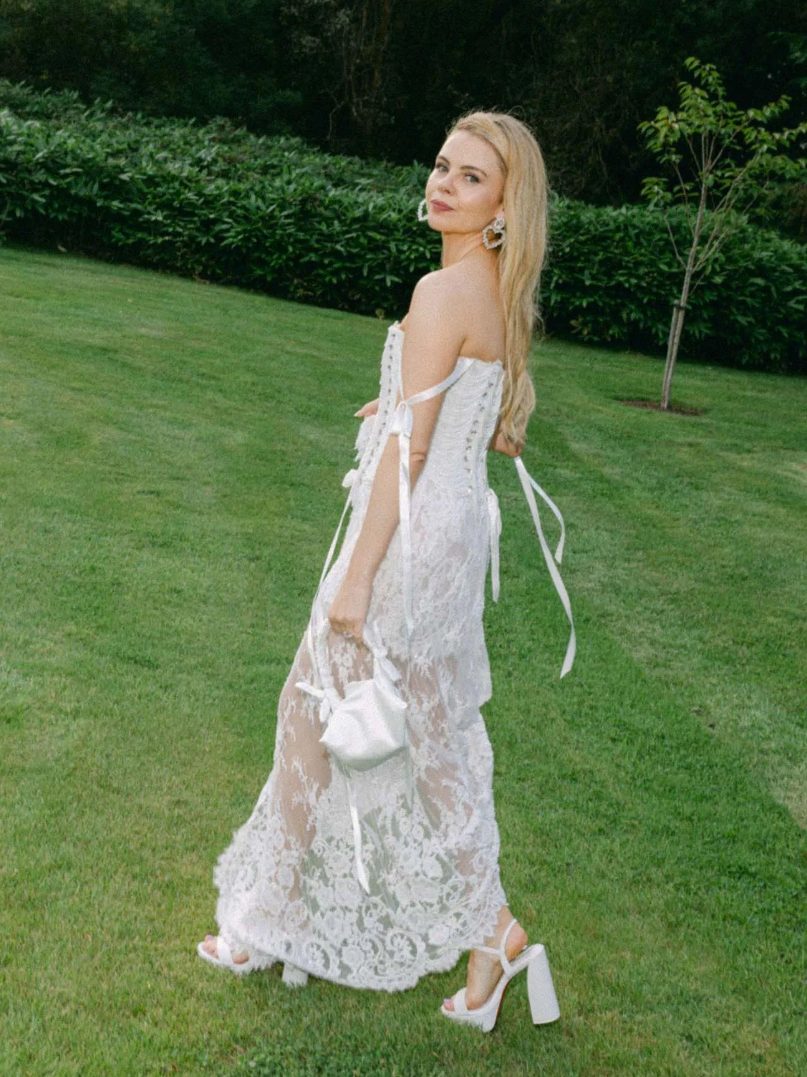Saoirse-Monica Jackson in a white lace wedding dress holding a small ivory satin ruched handle handbag standing on grass with greenery in the background
