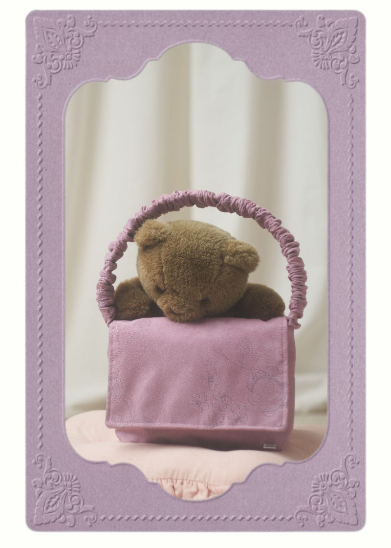 Teddy bear looking through the handle or a lilac handbag, sat on a pink cushion. Set on a white background