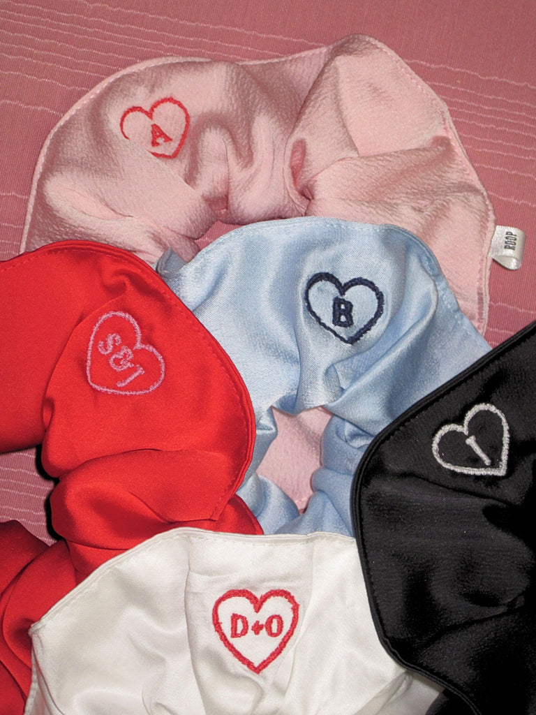 Five satin scrunchies embroidered with hearts that contain initials, set on a pink moire fabric background
