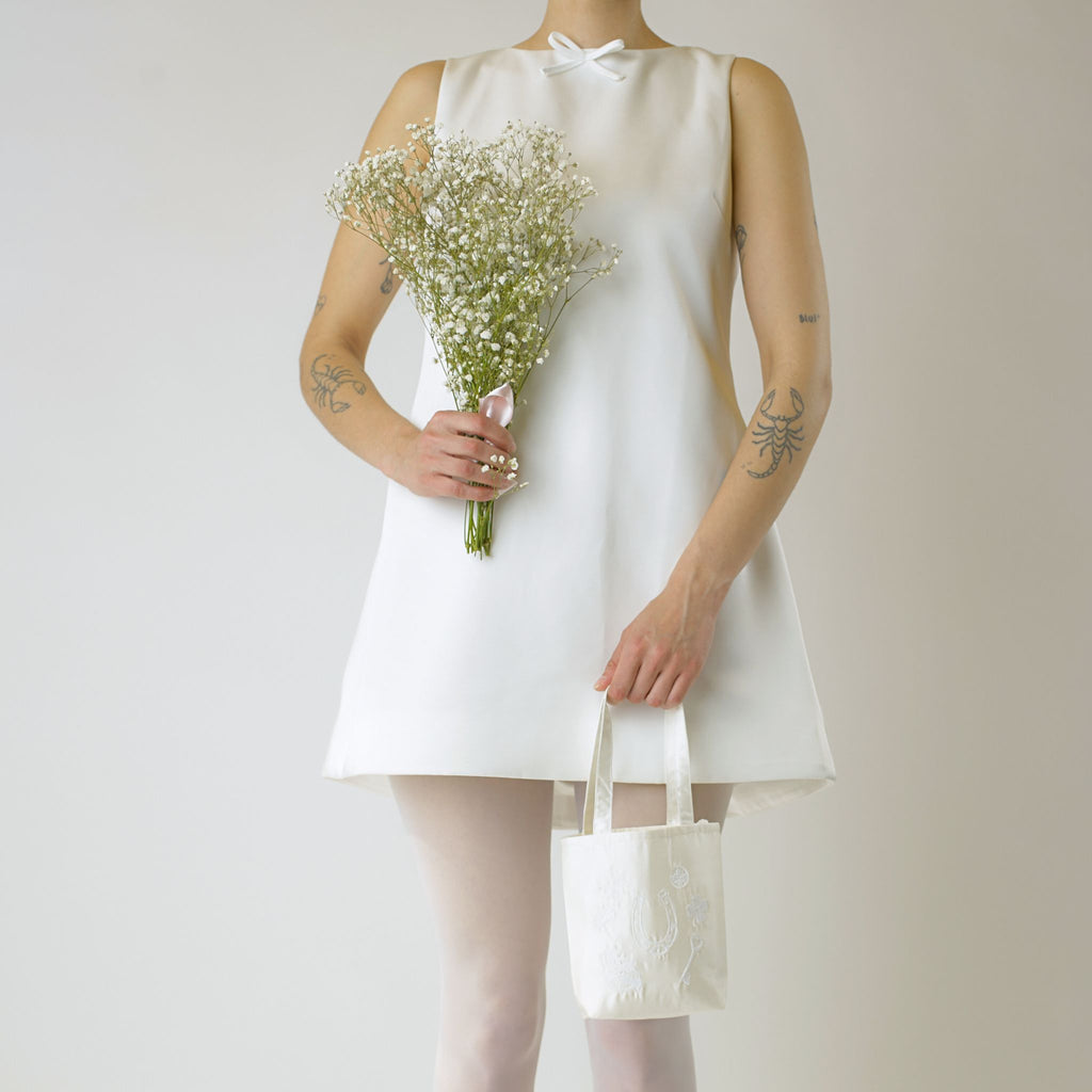 Person wearing an ivory short wedding dress holding a bouquet of baby's breath and an ivory small satin tote bag on a plain background