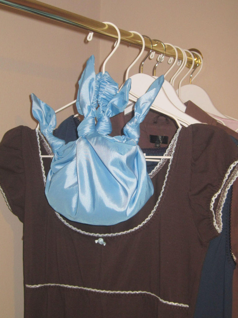 Blue taffeta knotted handbag on a white hanger with a brown dress snd other garments hanging on a gold rail. Beige wall.