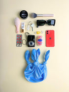 Collection of everyday items including a phone, camera, and makeup items on a beige background. A small blue knitted handbag sits at the bottom.