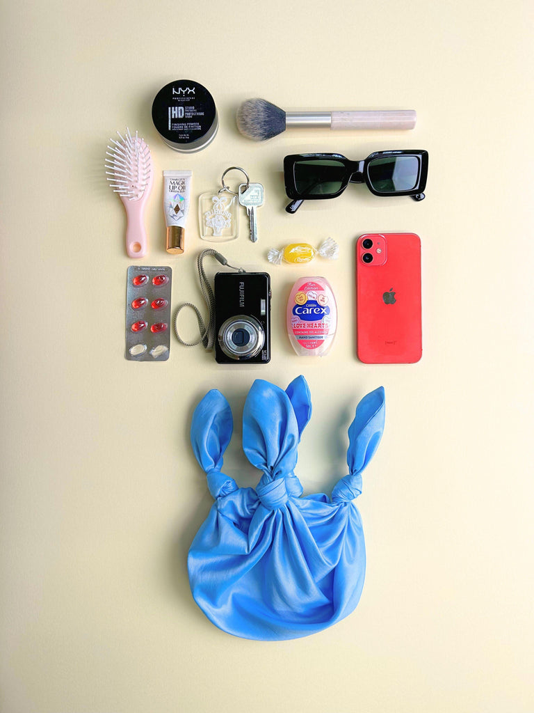 Collection of everyday items including a phone, camera, and makeup items on a beige background. A small blue knitted handbag sits at the bottom.