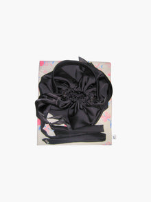 Black satin puffball drawstring handbag with its long straps coiled up, sat on top of a floral print fabric gift bag. Set on a white background.