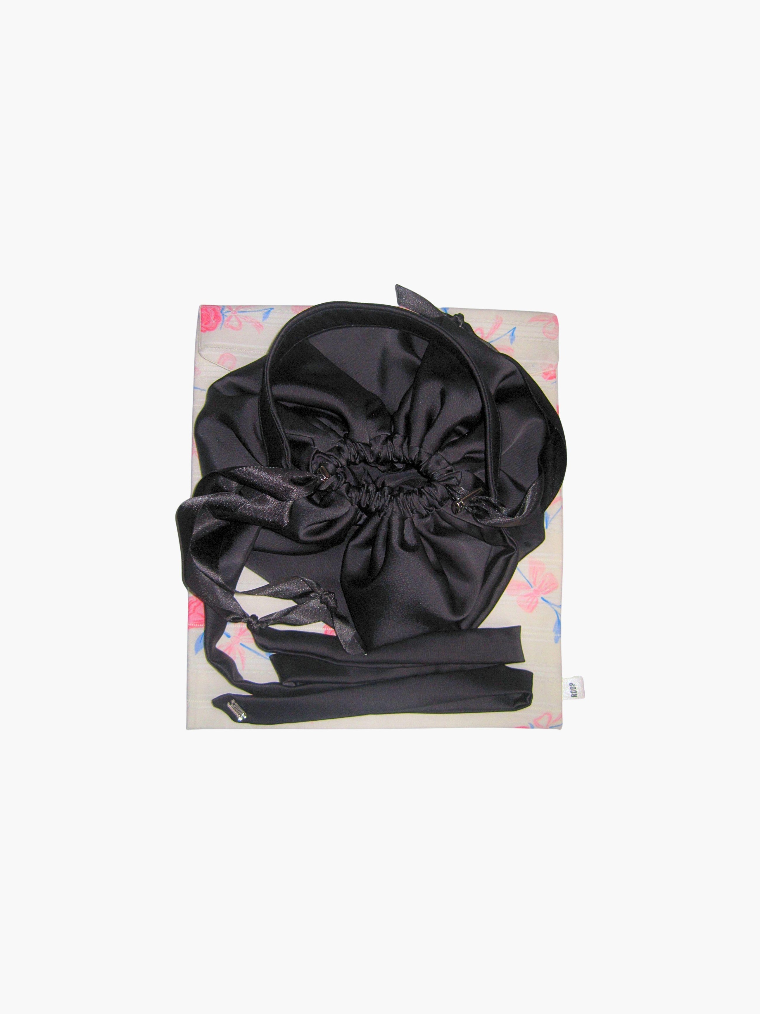 Black satin puffball drawstring handbag with its long straps coiled up, sat on top of a floral print fabric gift bag. Set on a white background.