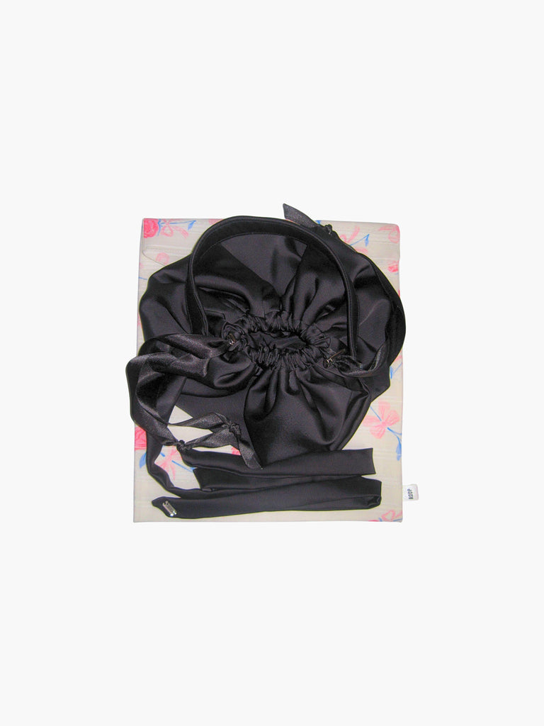 Black satin puffball drawstring handbag with its long straps coiled up, sat on top of a floral print fabric gift bag. Set on a white background.