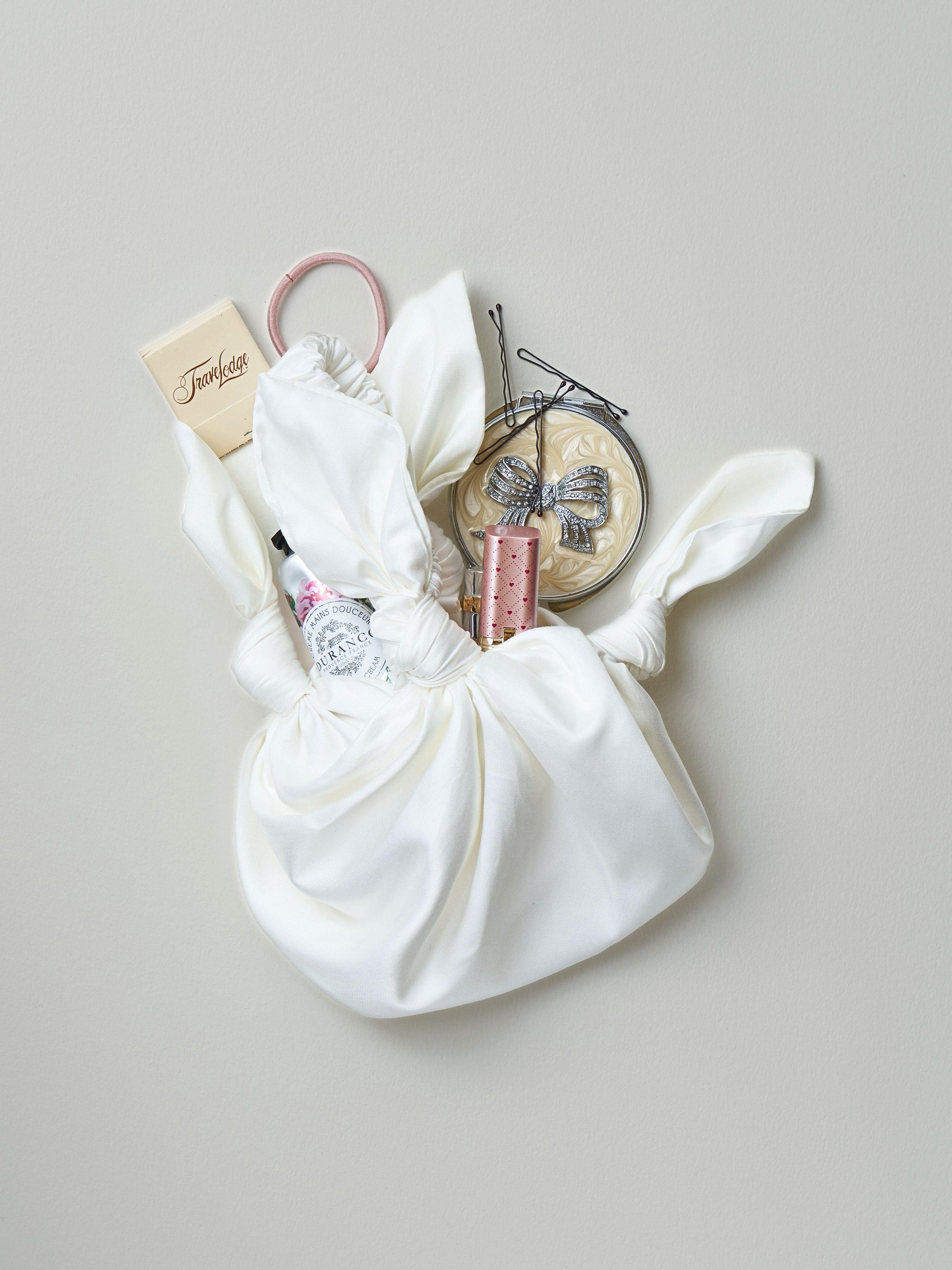 Ivory knotted satin handbag with possessions spilling out including a compact mirror, hair accessories, makeup and matches, sat on a light grey background