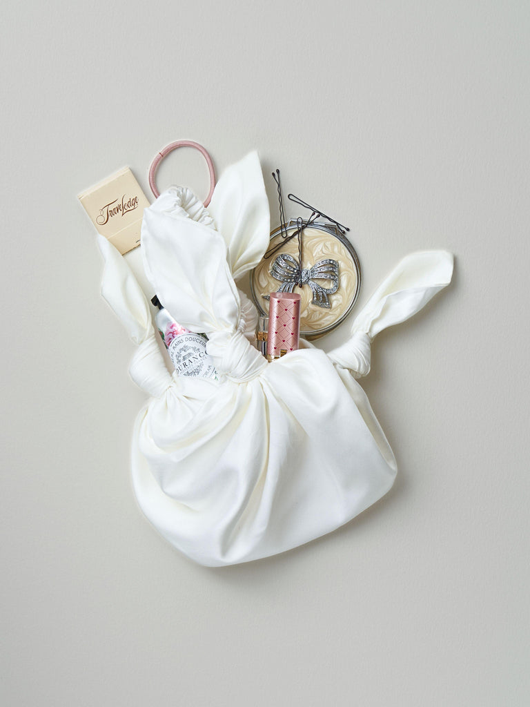 Ivory knotted satin handbag with possessions spilling out including a compact mirror, hair accessories, makeup and matches, sat on a light grey background