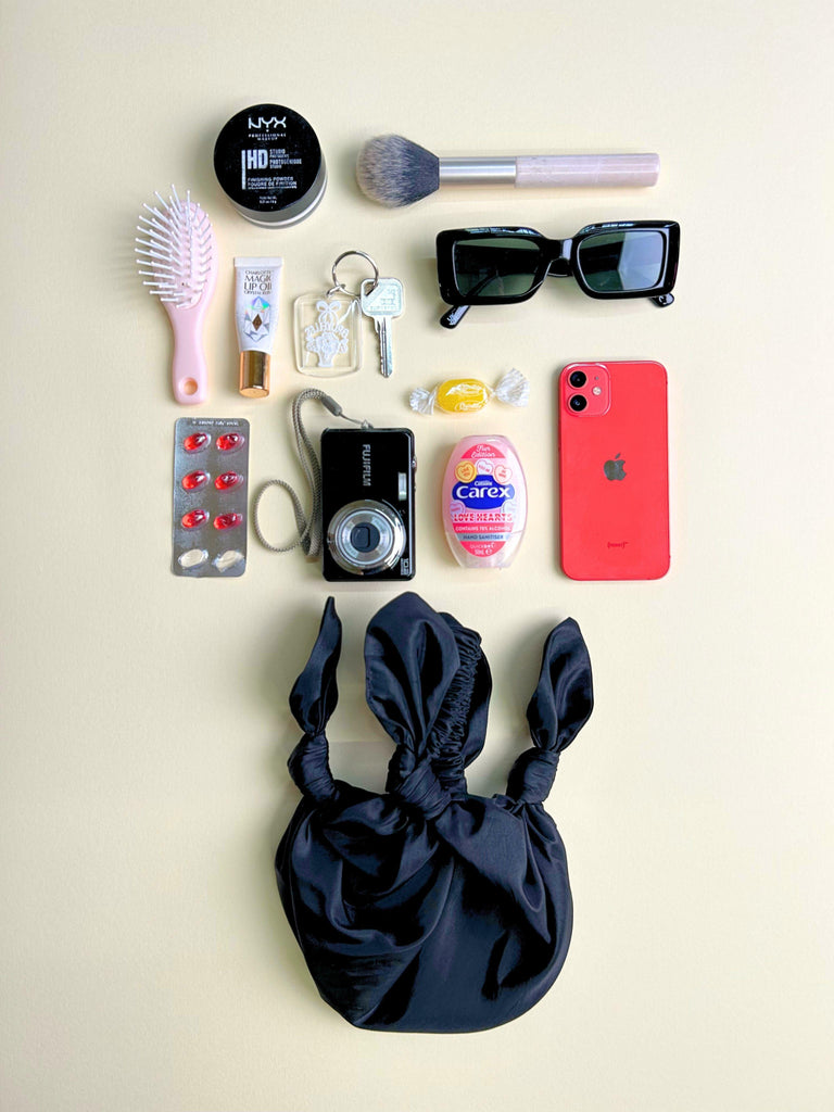 Collection of everyday items including a phone, makeup products, and personal care items on a beige background. Black knotted handbag sits at the bottom