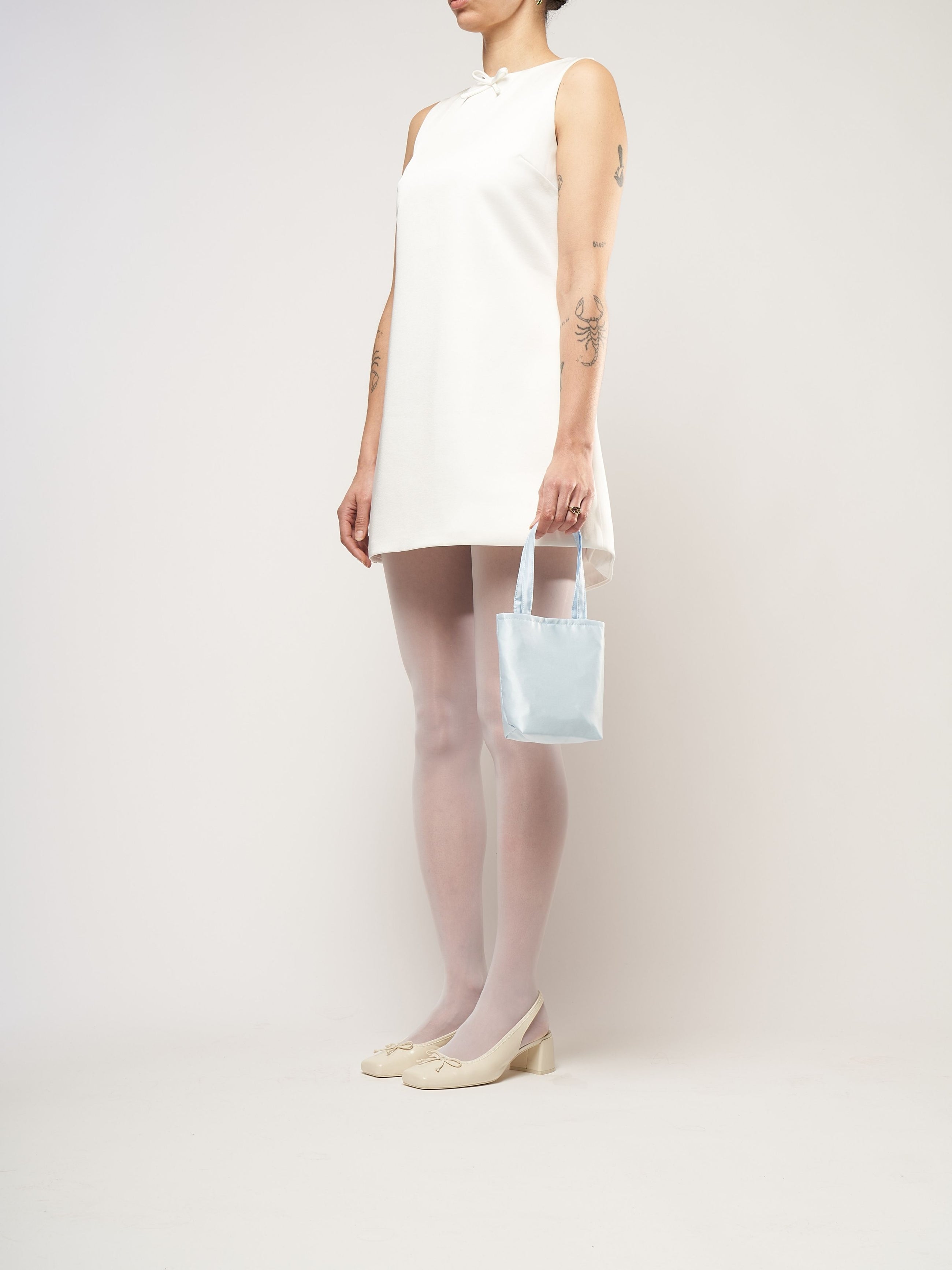 A person in a white sleeveless dress stands against a plain background. They hold a small light blue bag and wear white tights with cream heels.