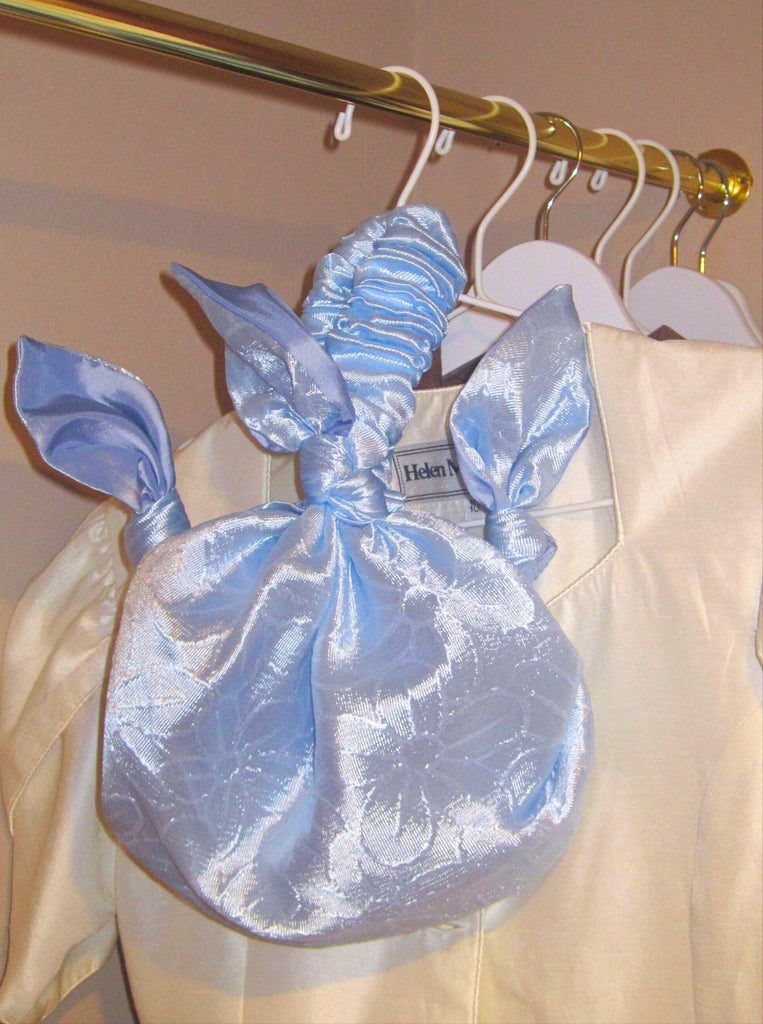 Light blue floral satin handbag hanging on a clothes rail with ivory garments in the back. Gold clothes rain, beige walls.
