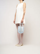 A person in a white sleeveless dress stands against a plain background. They hold a small light blue bag and wear white tights with cream heels.