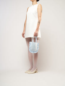 A person in a white sleeveless dress stands against a plain background. They hold a small light blue bag and wear white tights with cream heels.