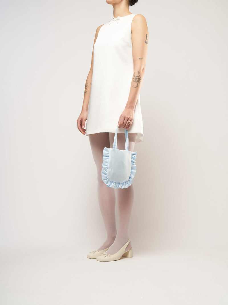 A person in a white sleeveless dress stands against a plain background. They hold a small light blue bag and wear white tights with cream heels.