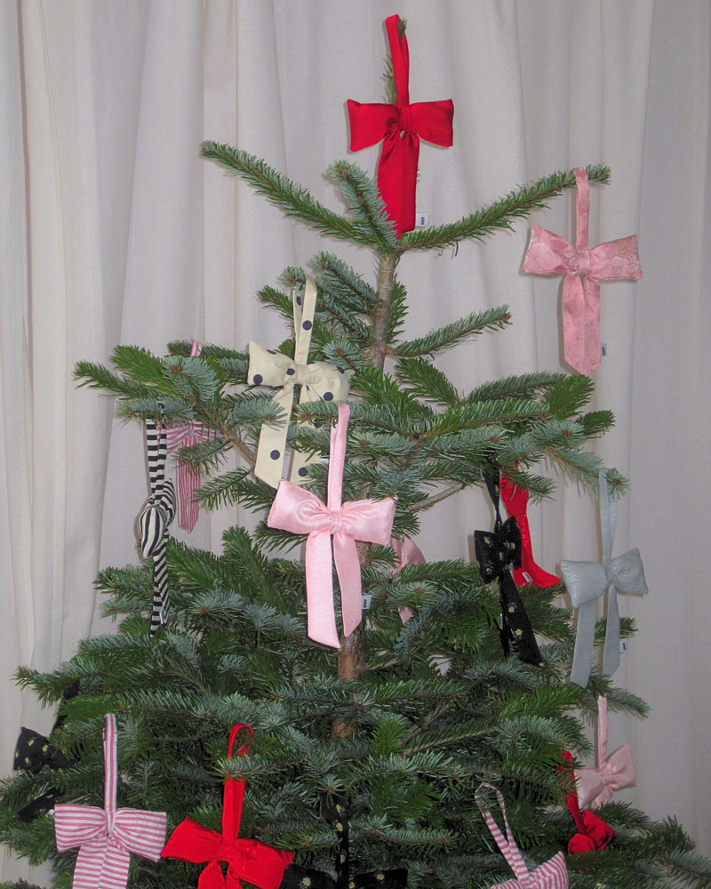 A Christmas tree adorned with colourful bows, including red, pink, polka dot, and striped patterns. The tree stands in front of white curtains.