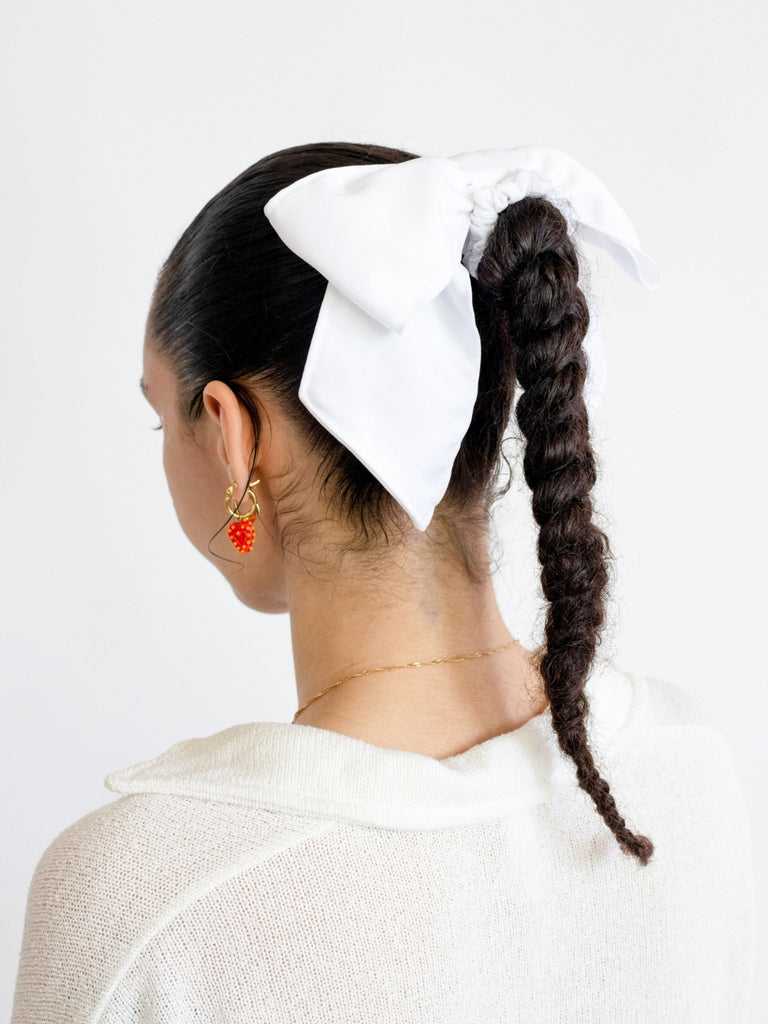 Person with a brown braid faces away, they have a large ivory bamboo silk satin hair bow scrunchie over their braid, set against a plain background.