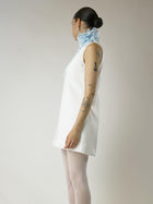 Person wearing a short ivory sleeveless wedding dress and sheer white stockings with a baby blue floral chiffon corsage around their neck, set on a plain background