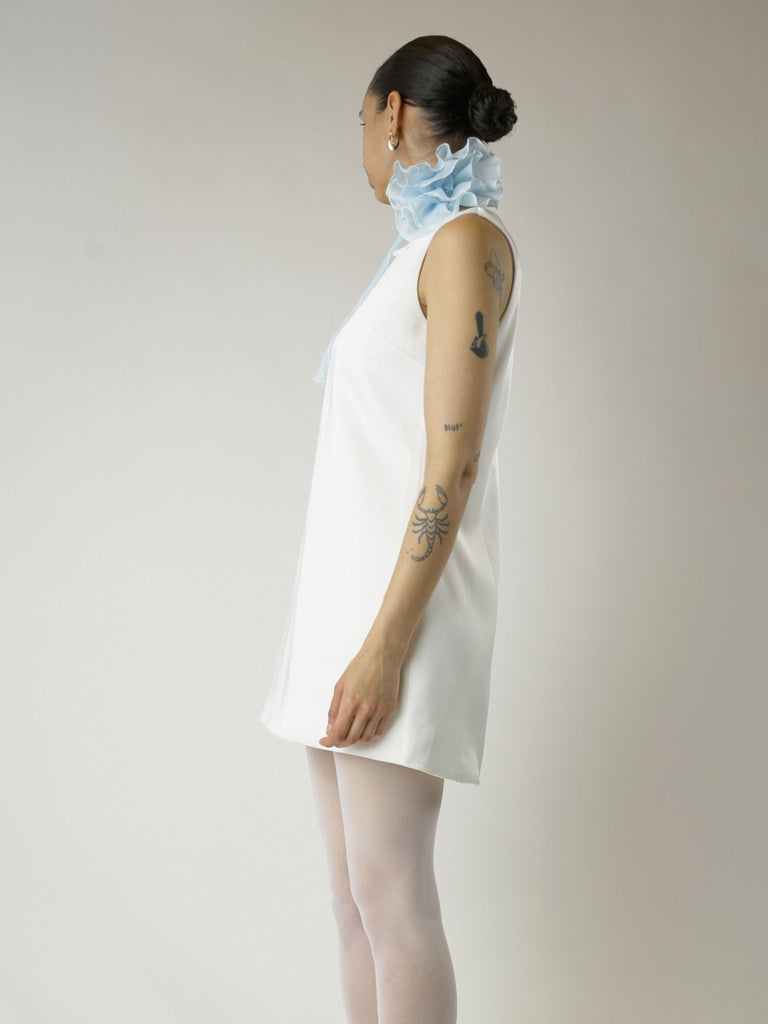 Person wearing a short ivory sleeveless wedding dress and sheer white stockings with a baby blue floral chiffon corsage around their neck, set on a plain background