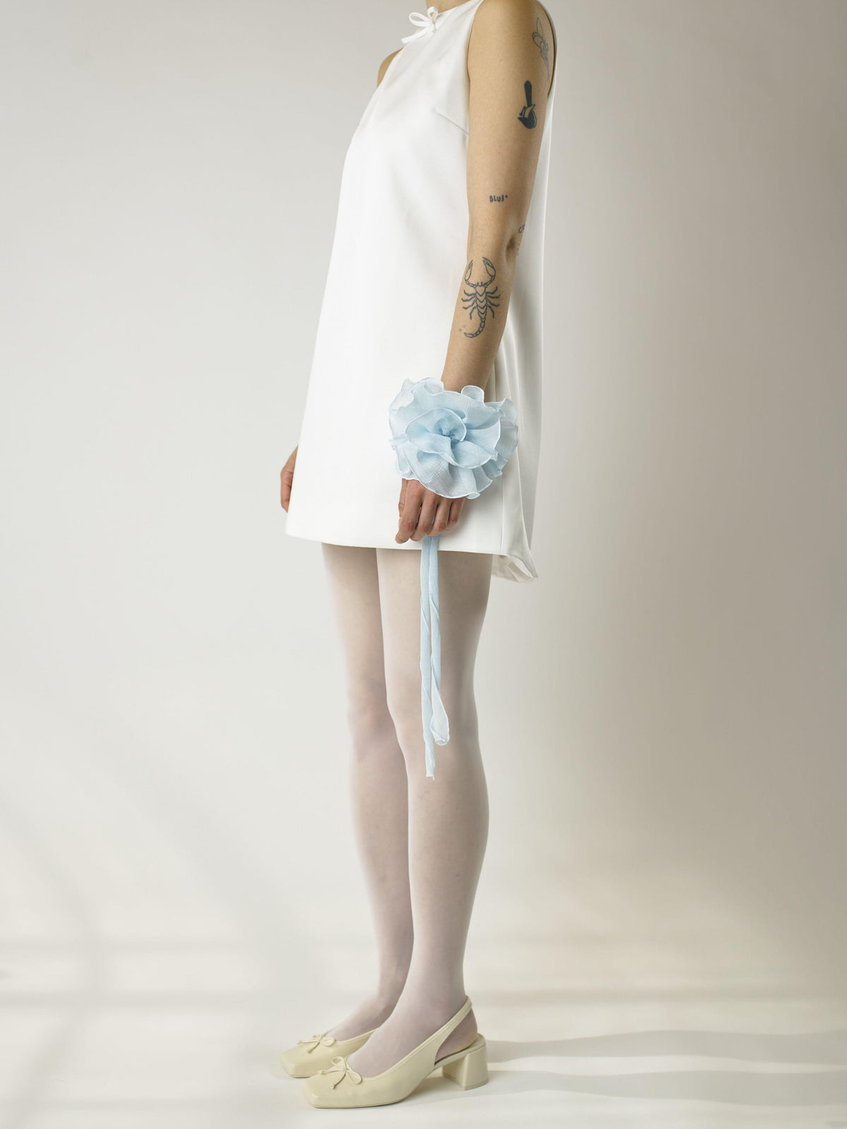 Person wearing a short ivory sleeveless wedding dress, sheer white stockings and heeled ivory ballet pumps, with a baby blue floral chiffon corsage around their wrist, set on a plain background