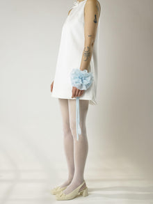 Person wearing a short ivory sleeveless wedding dress, sheer white stockings and heeled ivory ballet pumps, with a baby blue floral chiffon corsage around their wrist, set on a plain background