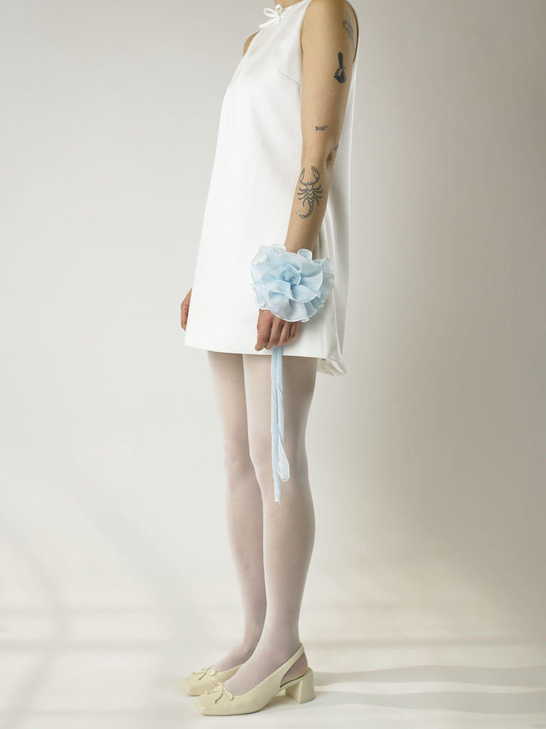 Person wearing a short ivory sleeveless wedding dress, sheer white stockings and heeled ivory ballet pumps, with a baby blue floral chiffon corsage around their wrist, set on a plain background