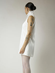 Person wearing a short ivory sleeveless wedding dress and sheer white stockings with a white floral chiffon corsage around their neck, set on a plain background