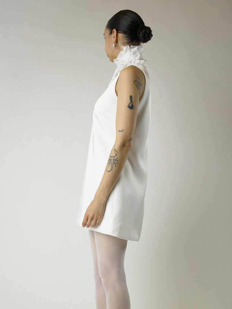 Person wearing a short ivory sleeveless wedding dress and sheer white stockings with a white floral chiffon corsage around their neck, set on a plain background