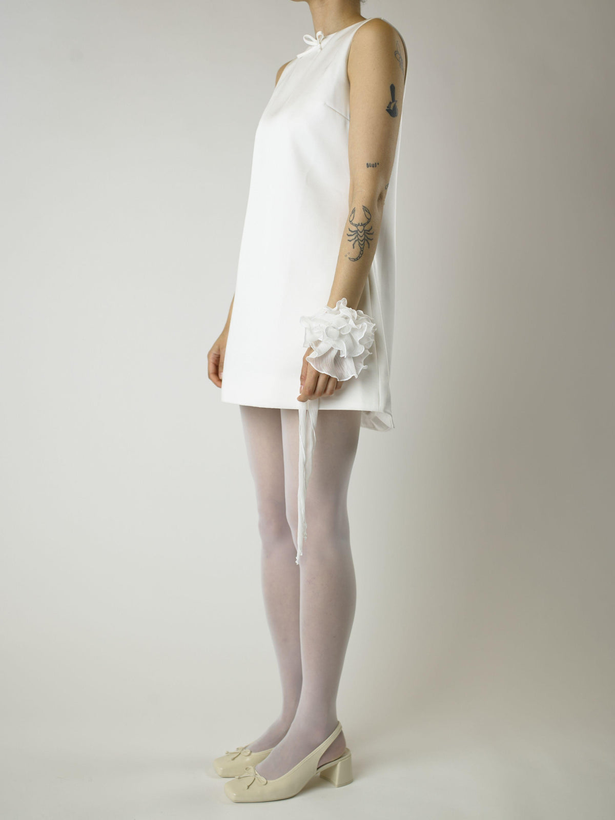 Person wearing a short ivory sleeveless wedding dress, sheer white stockings and heeled ivory ballet pumps, with a white floral chiffon corsage around their wrist, set on a plain background