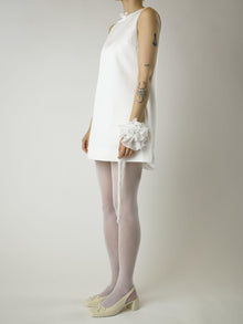 Person wearing a short ivory sleeveless wedding dress, sheer white stockings and heeled ivory ballet pumps, with a white floral chiffon corsage around their wrist, set on a plain background