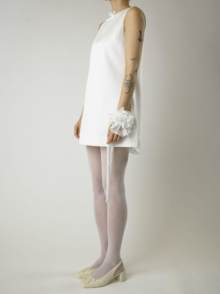 Person wearing a short ivory sleeveless wedding dress, sheer white stockings and heeled ivory ballet pumps, with a white floral chiffon corsage around their wrist, set on a plain background