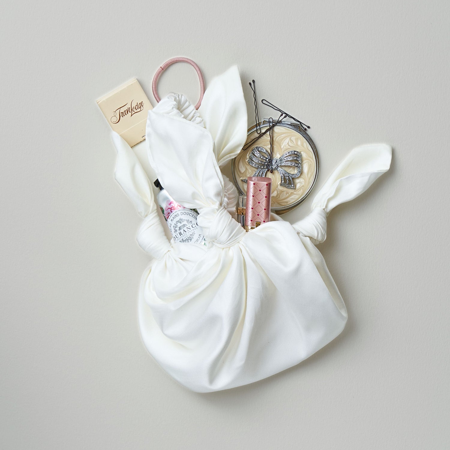 Ivory knotted small handbag with various small cosmetic items spilling out on a light grey background