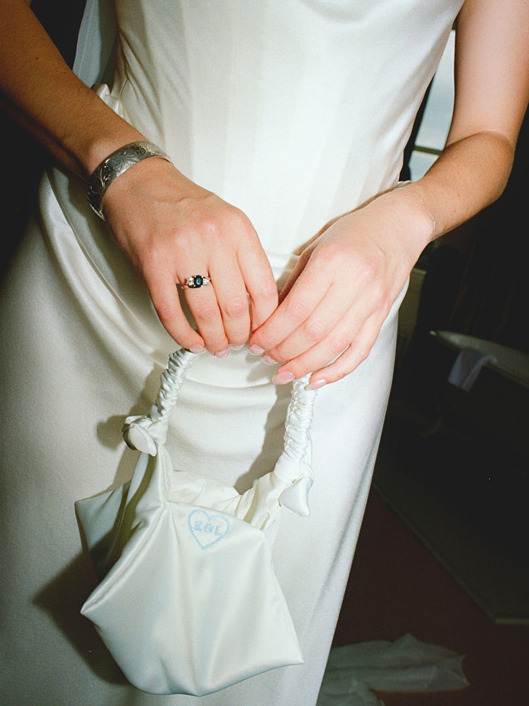 Person in a white wedding dress holding a small ivory satin ruched handle handbag embroidery with a blue heart with E&L inside.