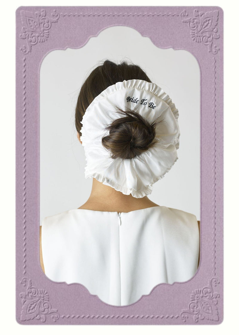 White scrunchie with 'Bride To Be' text embroidered on, worn around a brown haired bun. Image is inside a decorative purple frame