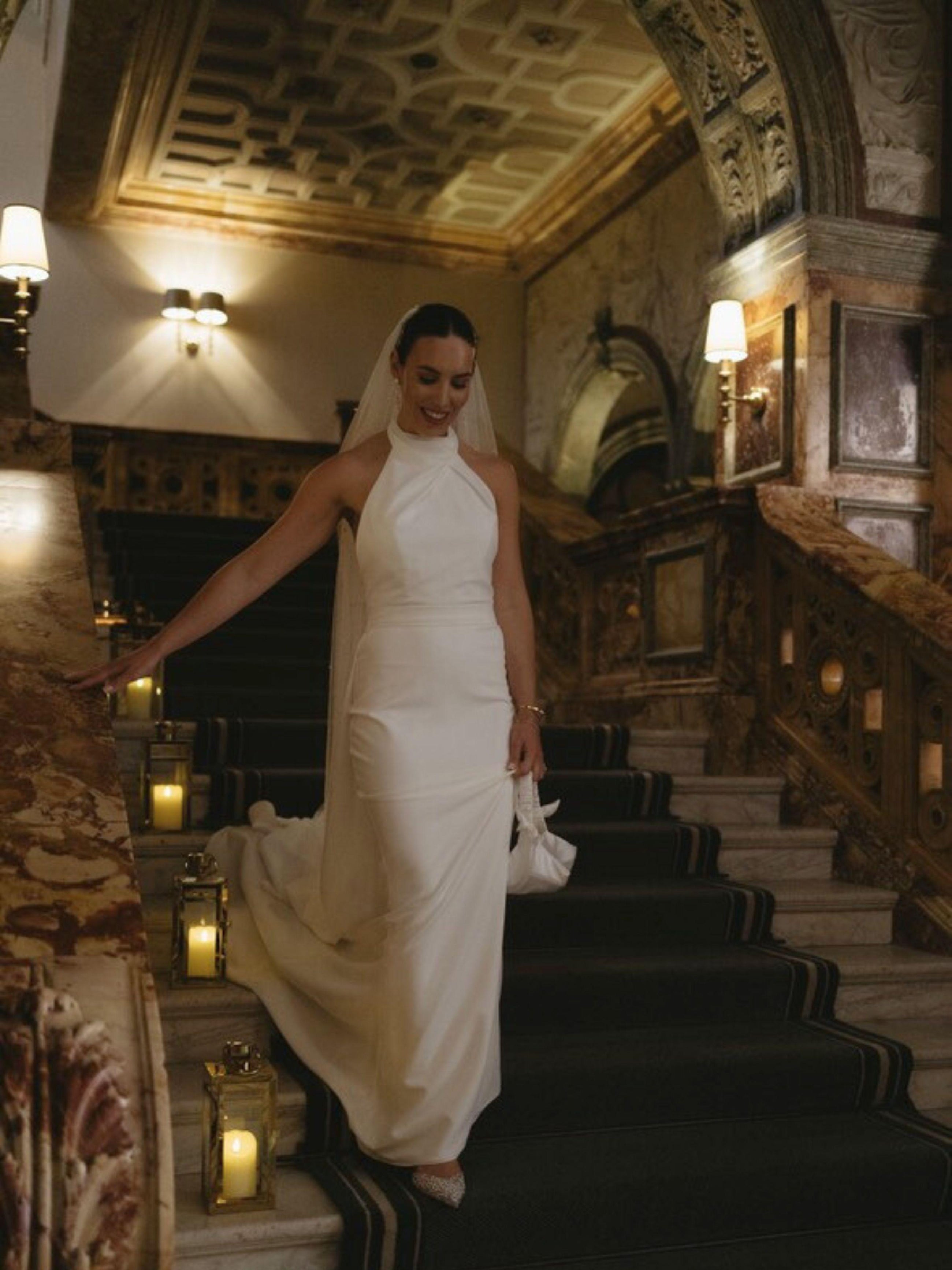 Woman in a white wedding dress standing on a staircase in an elegant interior setting. She holds a small ivory satin ruched handle handbag
