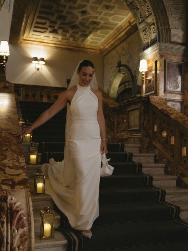 Woman in a white wedding dress standing on a staircase in an elegant interior setting. She holds a small ivory satin ruched handle handbag