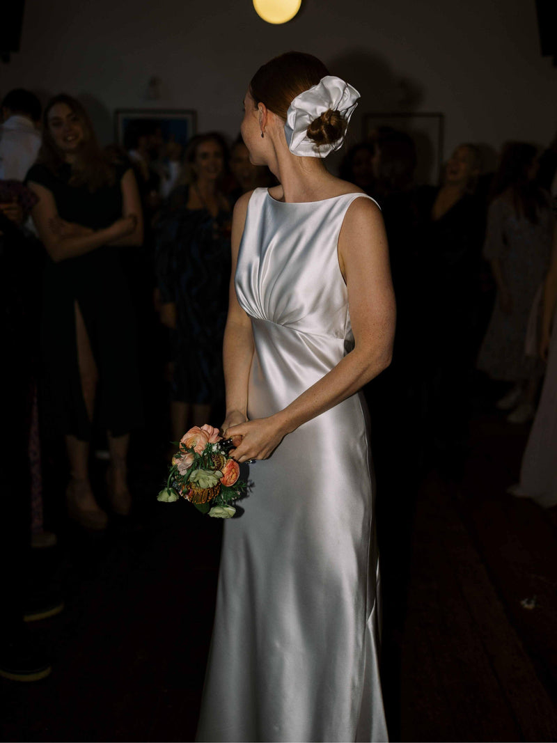 Woman in an ivory wedding dress wears a large ivory scrunchie over her bun, she's holding a bouquet in a dimly lit room with people in the background
