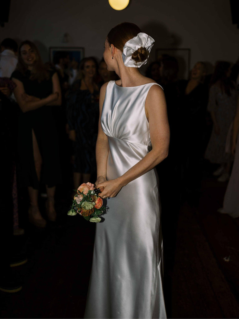 Woman in an ivory wedding dress wears a large ivory scrunchie over her bun, she's holding a bouquet in a dimly lit room with people in the background