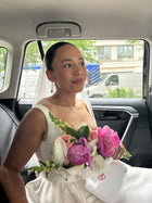 Bride in an ivory wedding dress  and veil sits in a car, she is holding a bouquet of flowers and an ivory satin box bag with a red heart embroidered on it.
