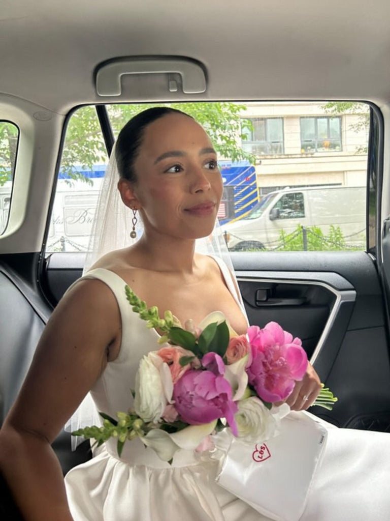 Bride in an ivory wedding dress  and veil sits in a car, she is holding a bouquet of flowers and an ivory satin box bag with a red heart embroidered on it.