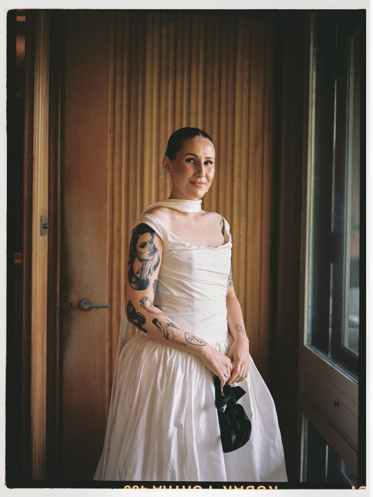 Woman in a white wedding dress and shawl, holds a black knotted handbag in front of her. She's standing in a room with a window and door.