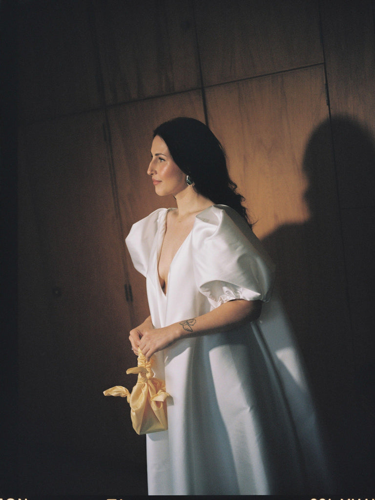 Woman in a white wedding dress holding a yellow bag against a wooden wall.