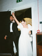 bride and groom enter a room happily. They are holding hands in the air, she holds a white satin knotted handbag