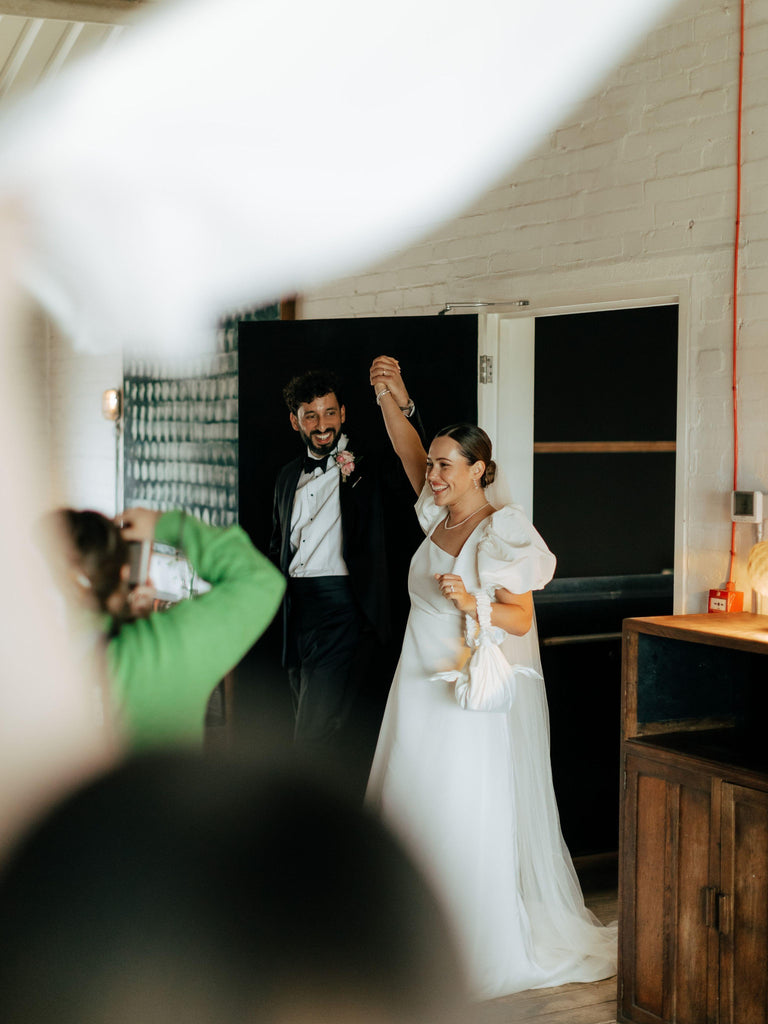 bride and groom enter a room happily. They are holding hands in the air, she holds a white satin knotted handbag