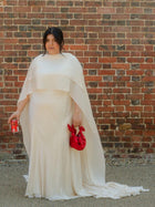 Person wearing an ivory wedding dress with a cape holding red knotted handbag and a can of coke zero against a brick wall.