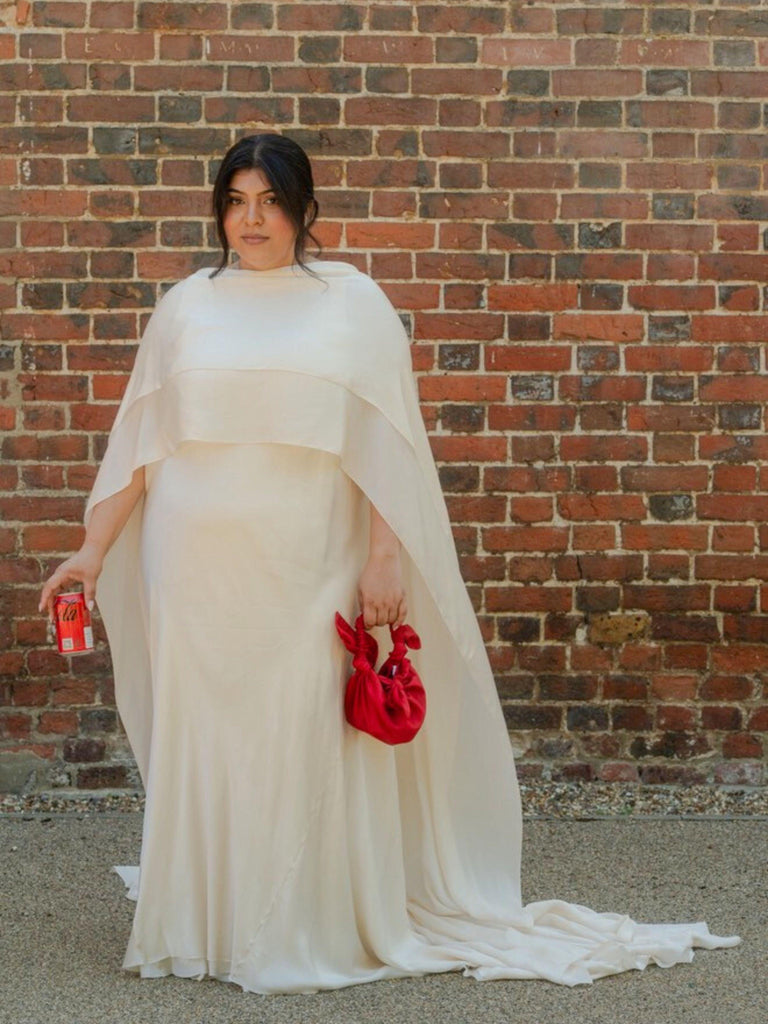 Person wearing an ivory wedding dress with a cape holding red knotted handbag and a can of coke zero against a brick wall.